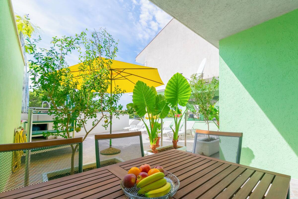 Taula de fusta amb un plat de fruita, cadires d'exterior, un para-sol groc i plantes en testos.
