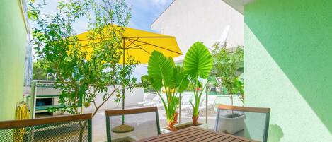 Une table en bois avec un panier de fruits, un parasol jaune et des plantes en pot dans un espace extérieur aménagé pour s’asseoir.