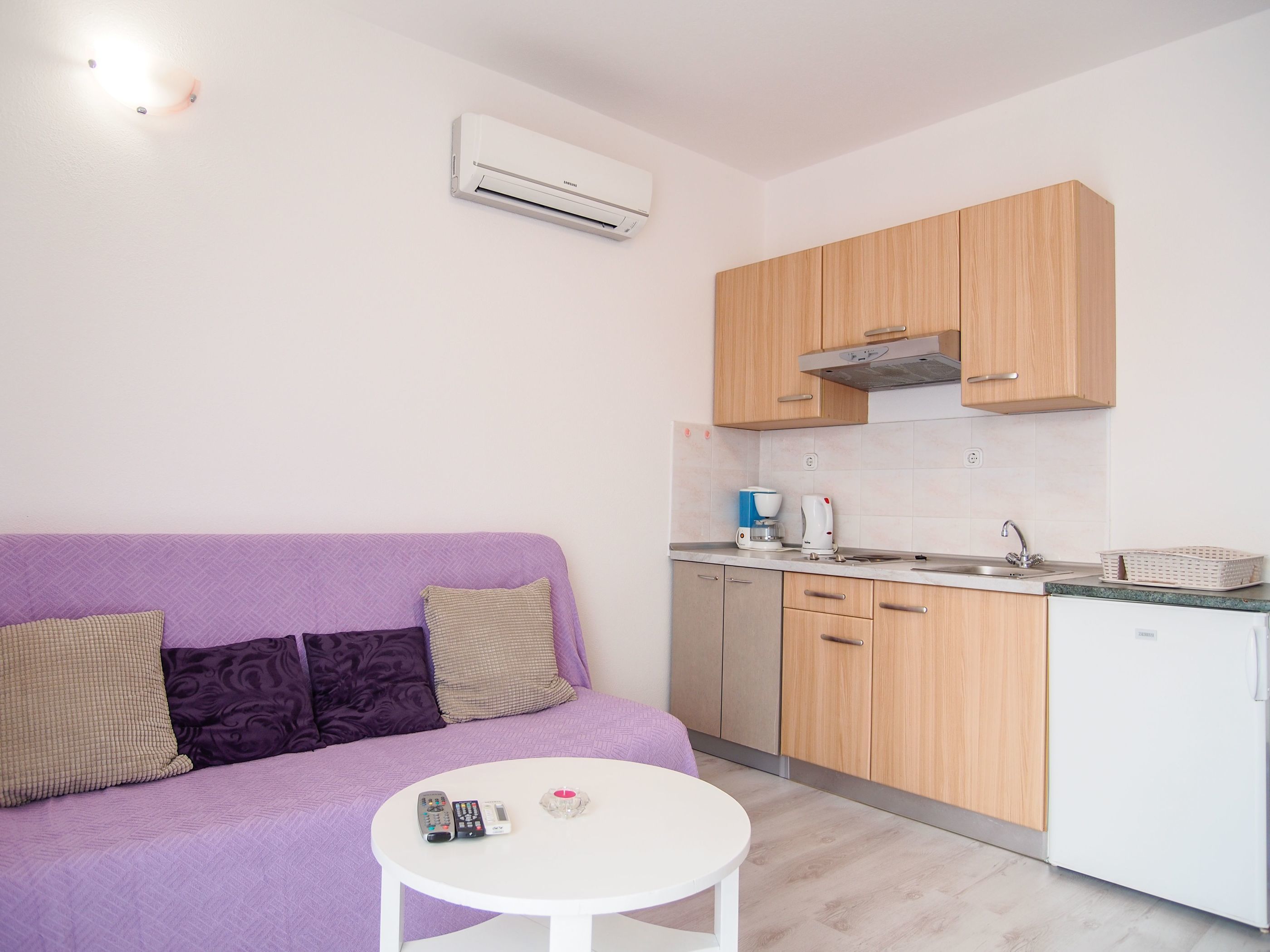 Un espace de vie compact comprenant un canapé violet, une petite table ronde, une kitchenette avec des meubles en bois et un réfrigérateur blanc.
