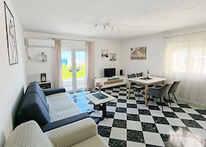 A living room with a sofa, a dining table with chairs, a TV, and a balcony with curtains.