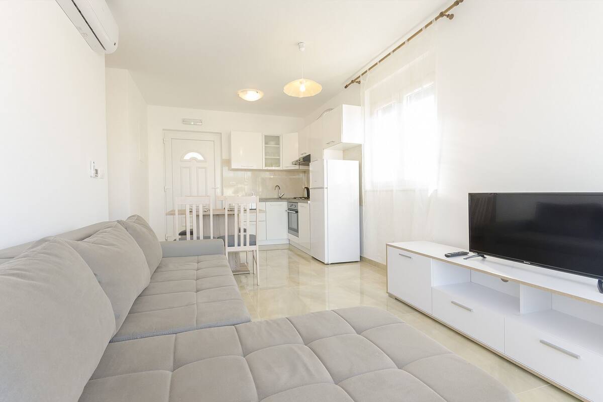 A modern living room with a grey sectional sofa, a flat-screen TV on a white stand, and a kitchen in the background.