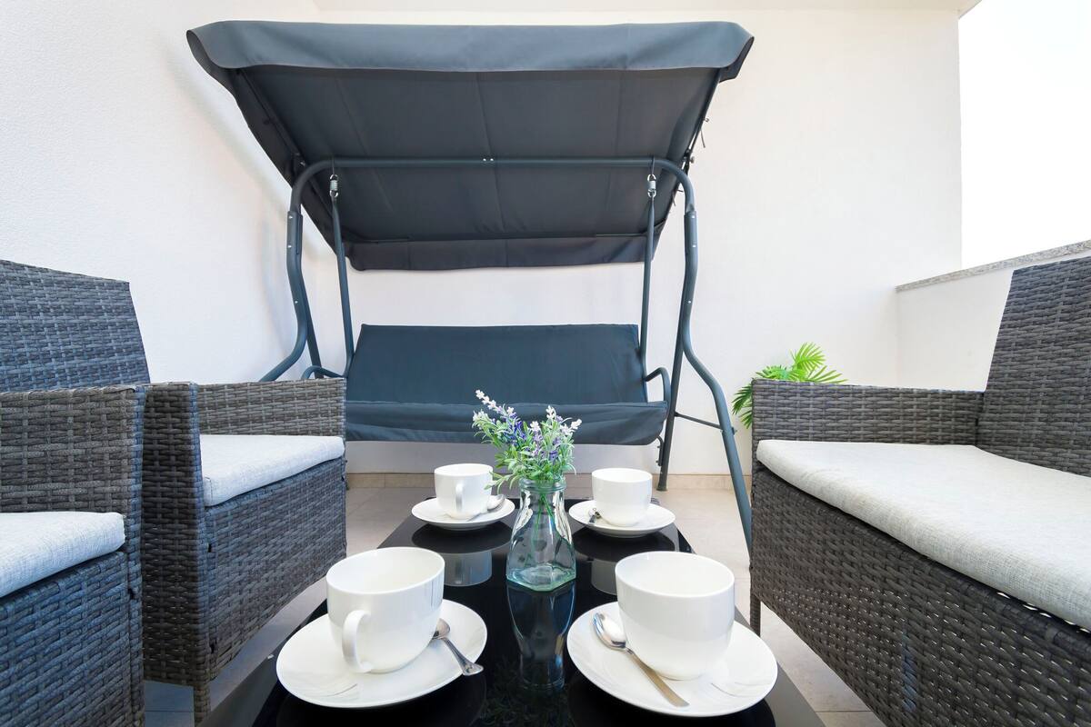 A dining area with wicker furniture, a glass table, white cups, and a small vase with flowers.
