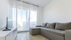 Ein modernes Wohnzimmer mit einem beige farbenen Sofa, einem Flachbildfernseher auf einem weißen Schreibtisch und leichten Vorhängen an einem großen Fenster.
