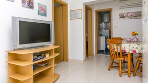 A living room with a TV on a wooden stand, a dining table with chairs, and a kitchen visible through an open door. - Apartment Bakovic (Rogoznica)