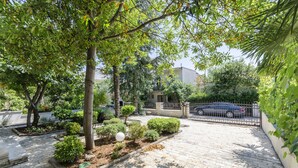 A paved courtyard with a variety of plants and trees, a gated entrance, and a parked car. - Apartments Karolina (Vodice)