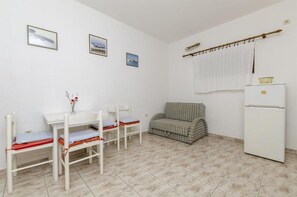 A small, clean room with a white table, four chairs, a patterned sofa, a white refrigerator, and three framed pictures on the wall. - Apartments Petra (Rogoznica)