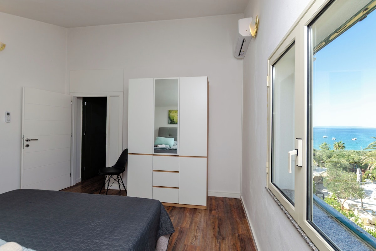 A bedroom with a bed, a wardrobe, a desk, and a large window with a view of the sea.