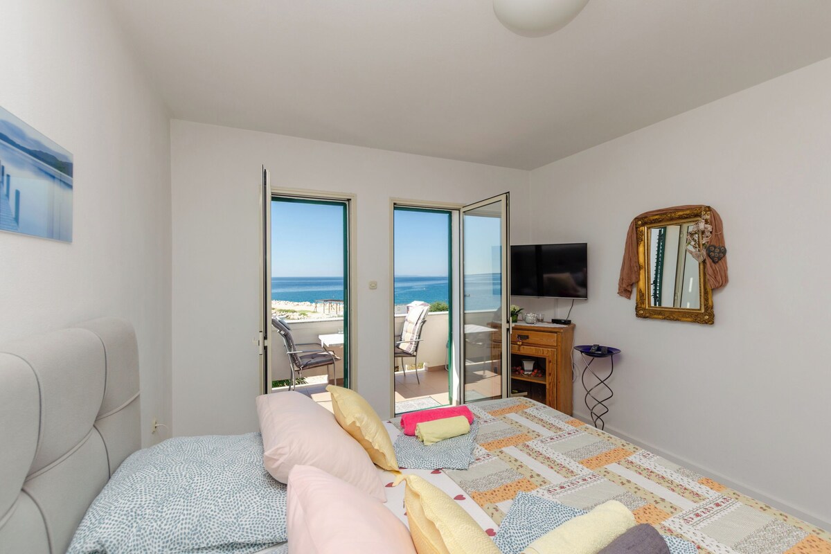A bedroom with a bed, a TV, a mirror, and a view of the sea through an open door.