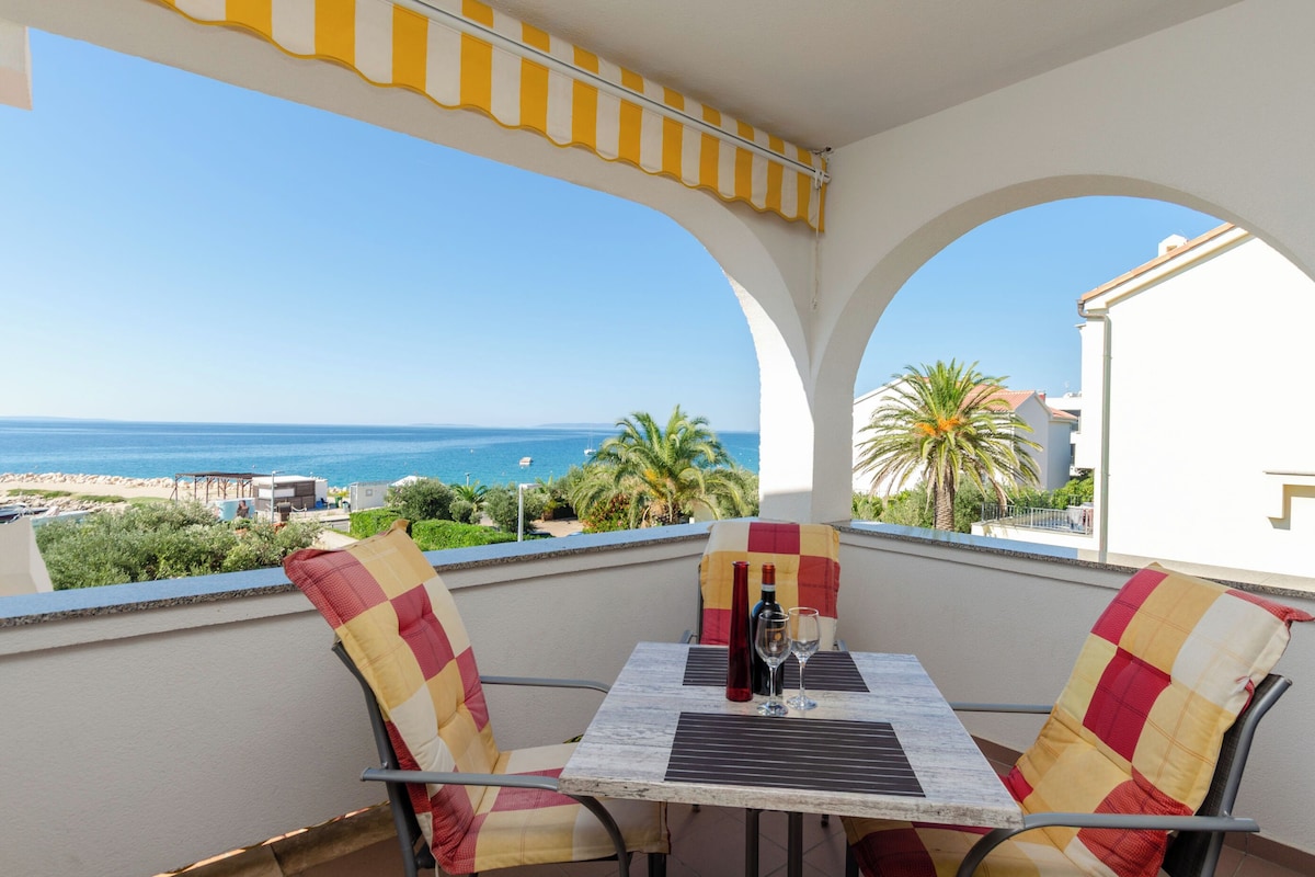 A balcony with a table and chairs, overlooking a beach with a clear view of the sea and distant buildings.