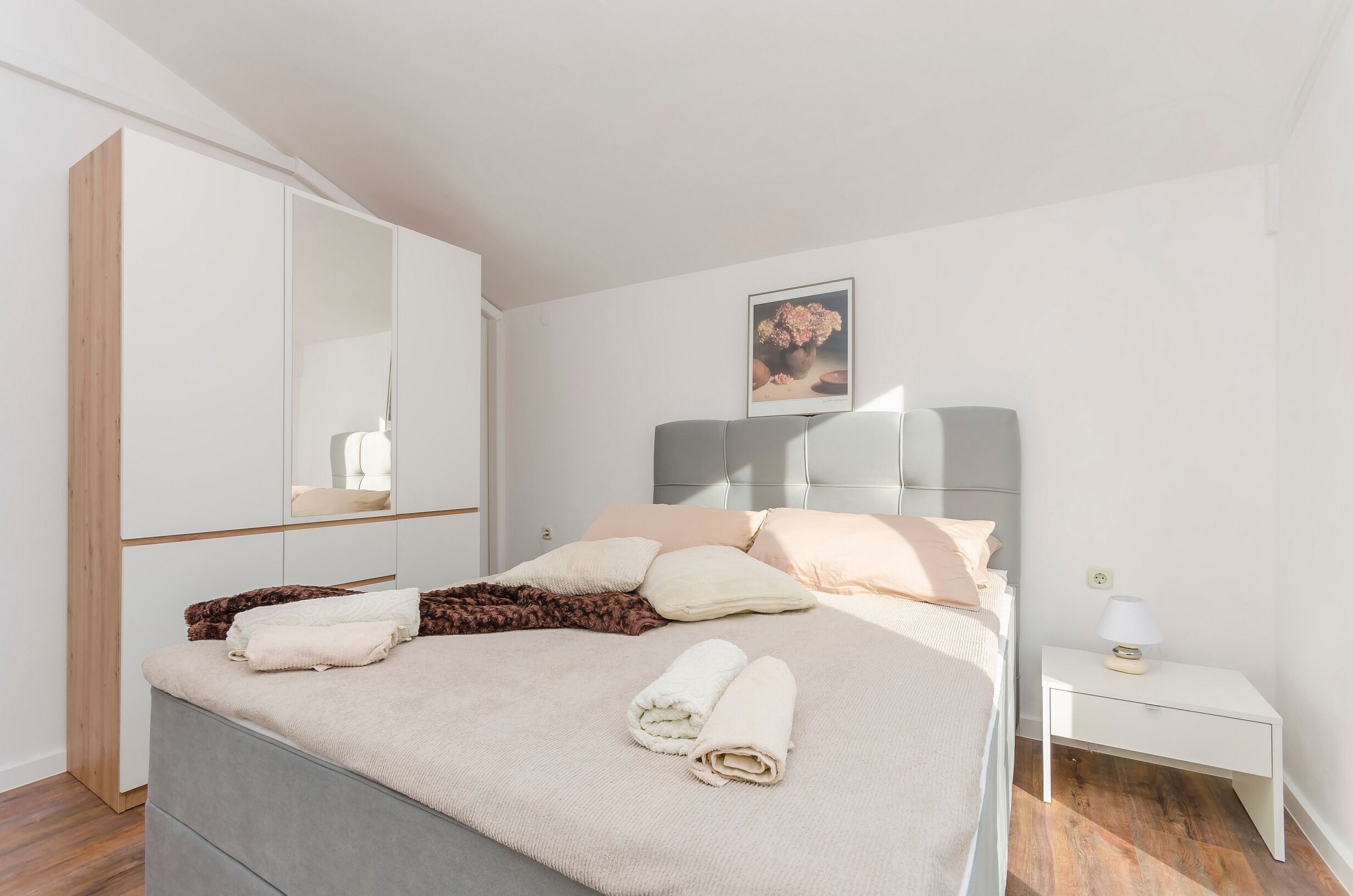 A neatly made bed with pillows and a blanket, a bedside table with a lamp, a mirror, and a framed picture on the wall.