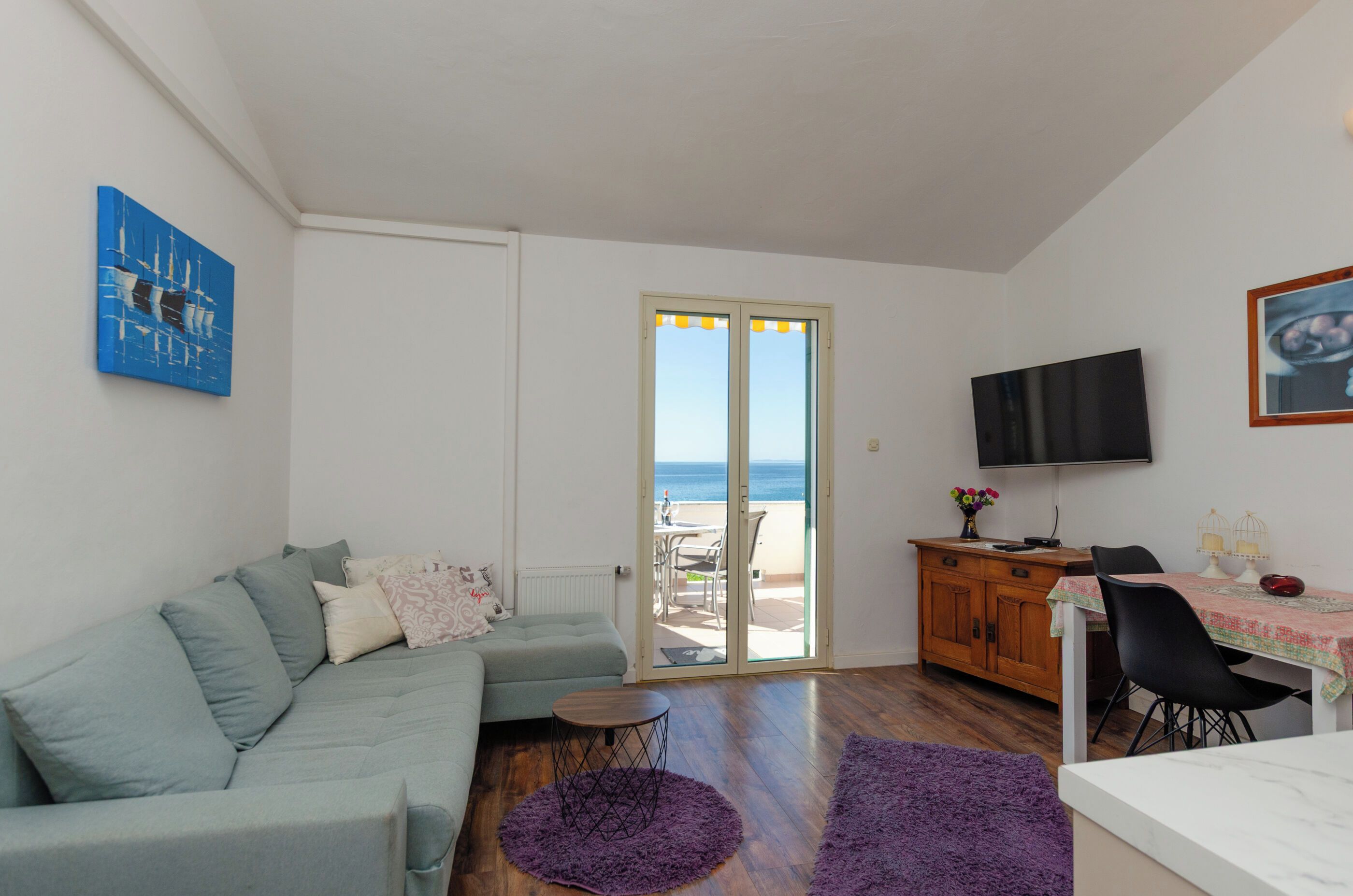A living room with a sofa, a small round table, a purple rug, a wooden cabinet, a TV, and a view of the sea through a glass door.