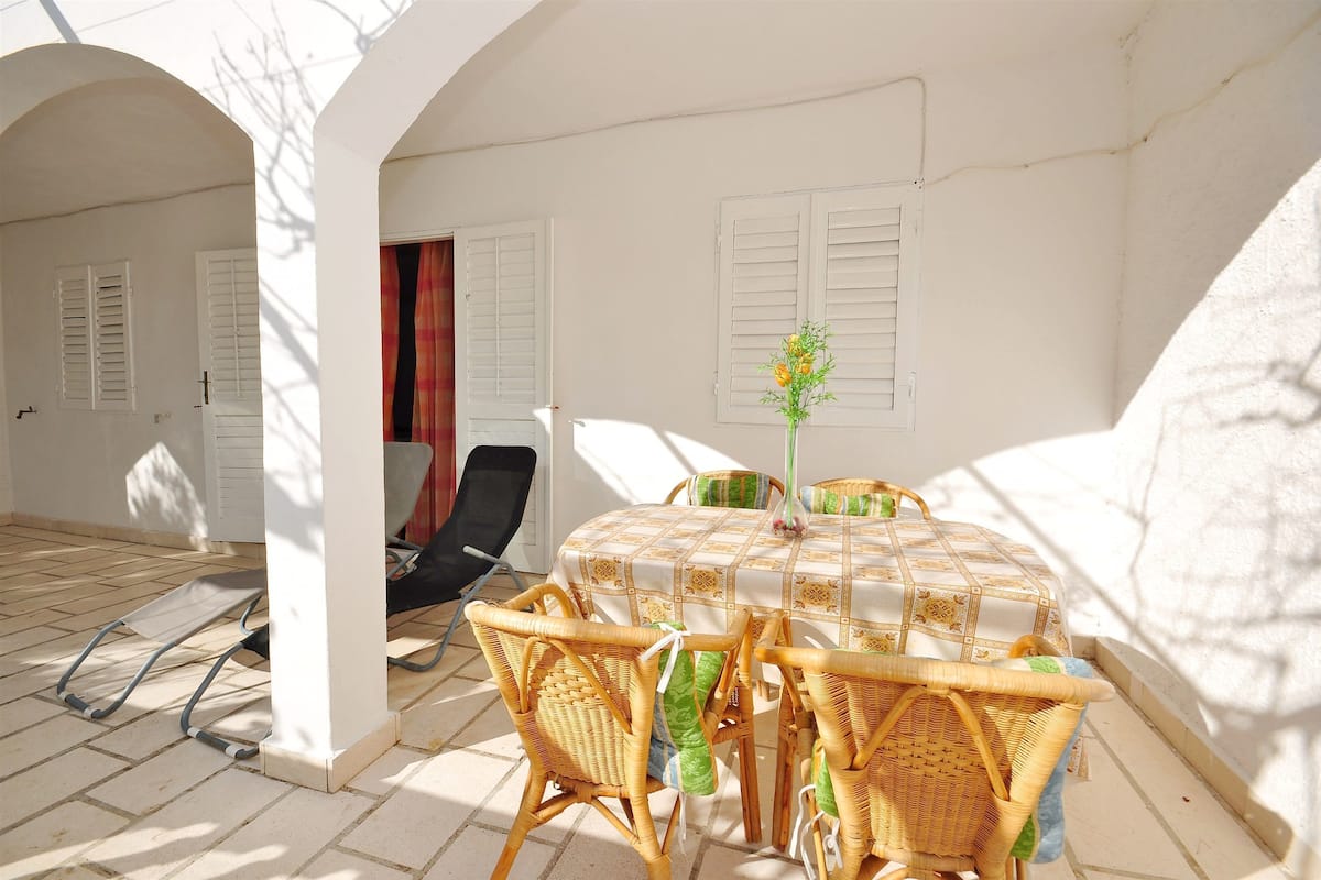 A sunlit patio with a round table and wicker chairs, white walls, and shutters.