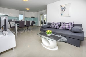 Un salon moderne avec un canapé gris, une table basse en verre et un coin repas comprenant une table et des chaises blanches.