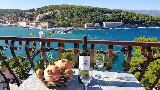 A table with a bottle of wine, a glass, and a basket of fruits on a balcony overlooking a coastal town. - Apartments and Room Caric (Jelsa)