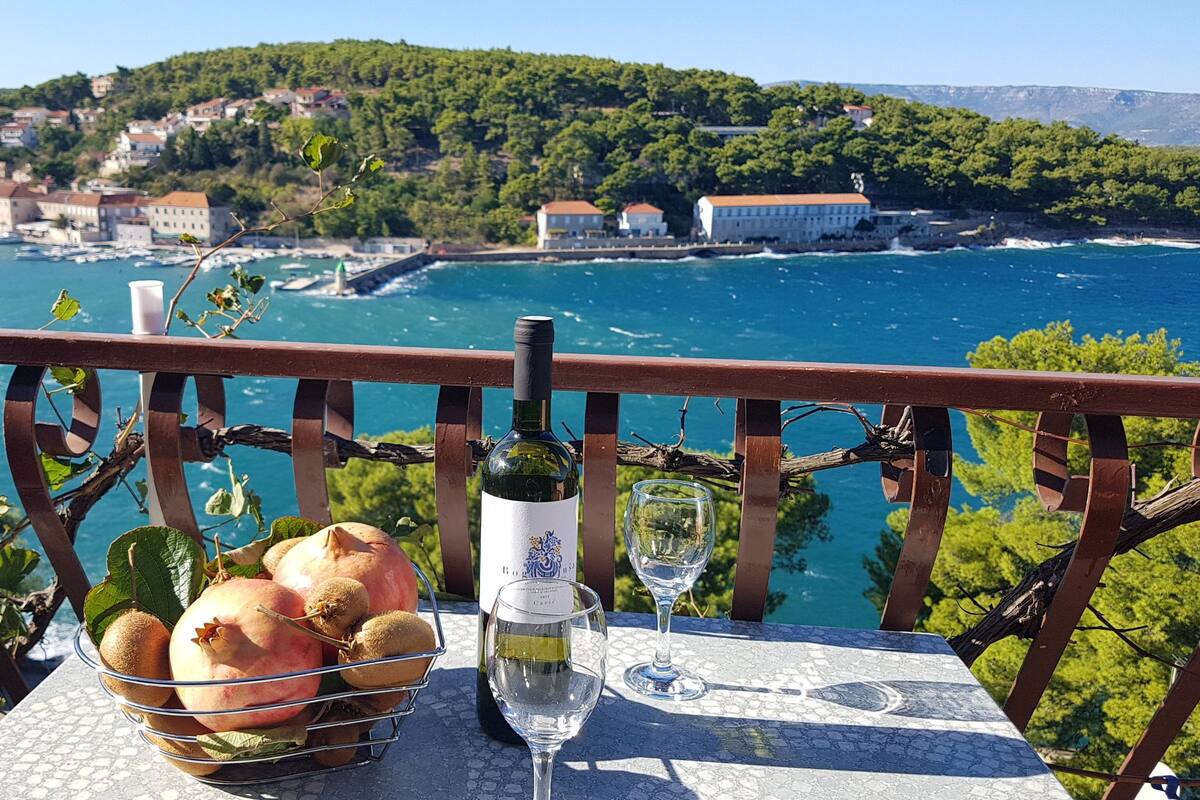Un tavolo con una bottiglia di vino, un bicchiere e un cesto di frutta su un balcone con vista su una cittadina costiera.