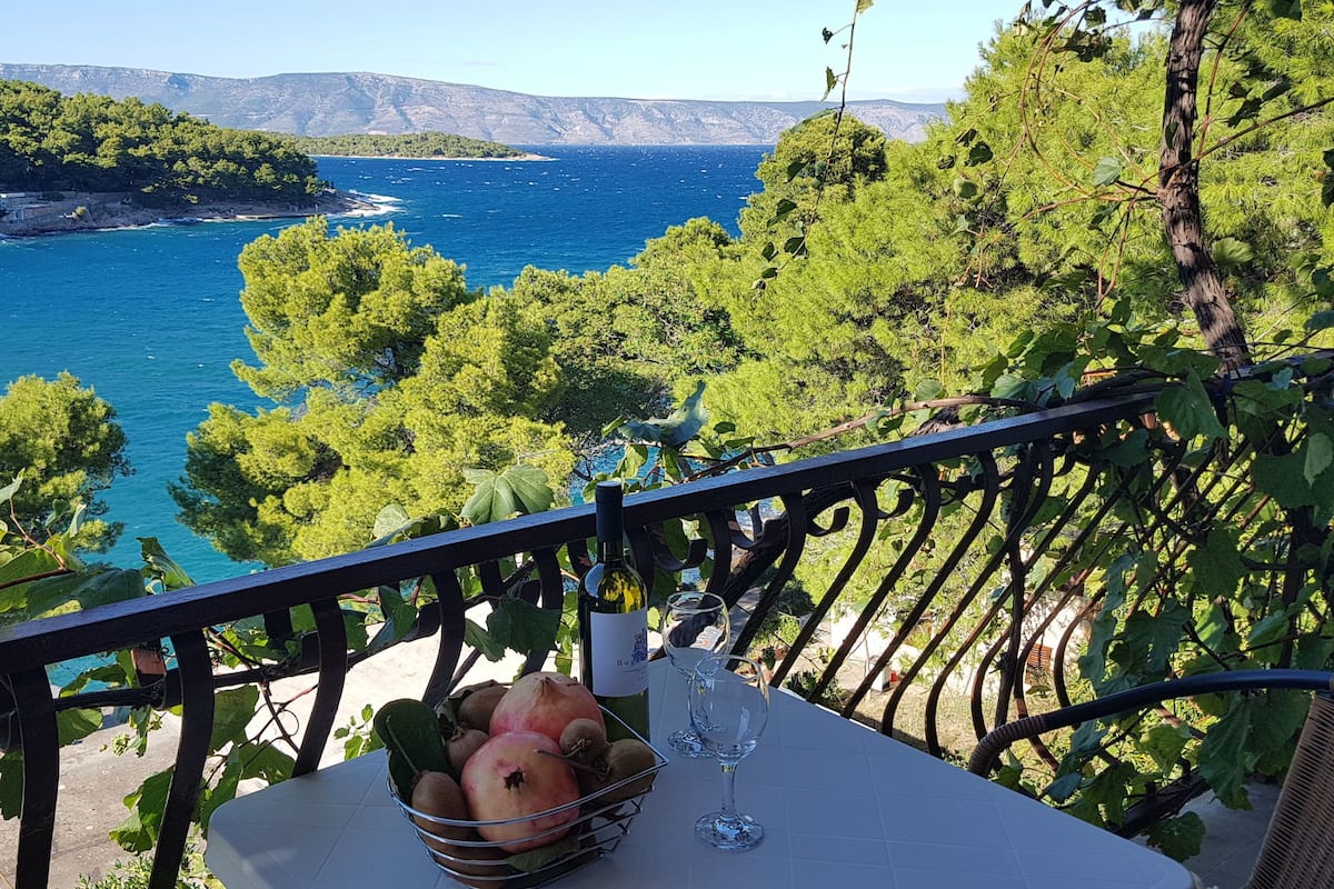 Un tavolo con una bottiglia di vino, un bicchiere e un cesto di frutta su un balcone affacciato su una vista costiera con alberi e una costa in lontananza.