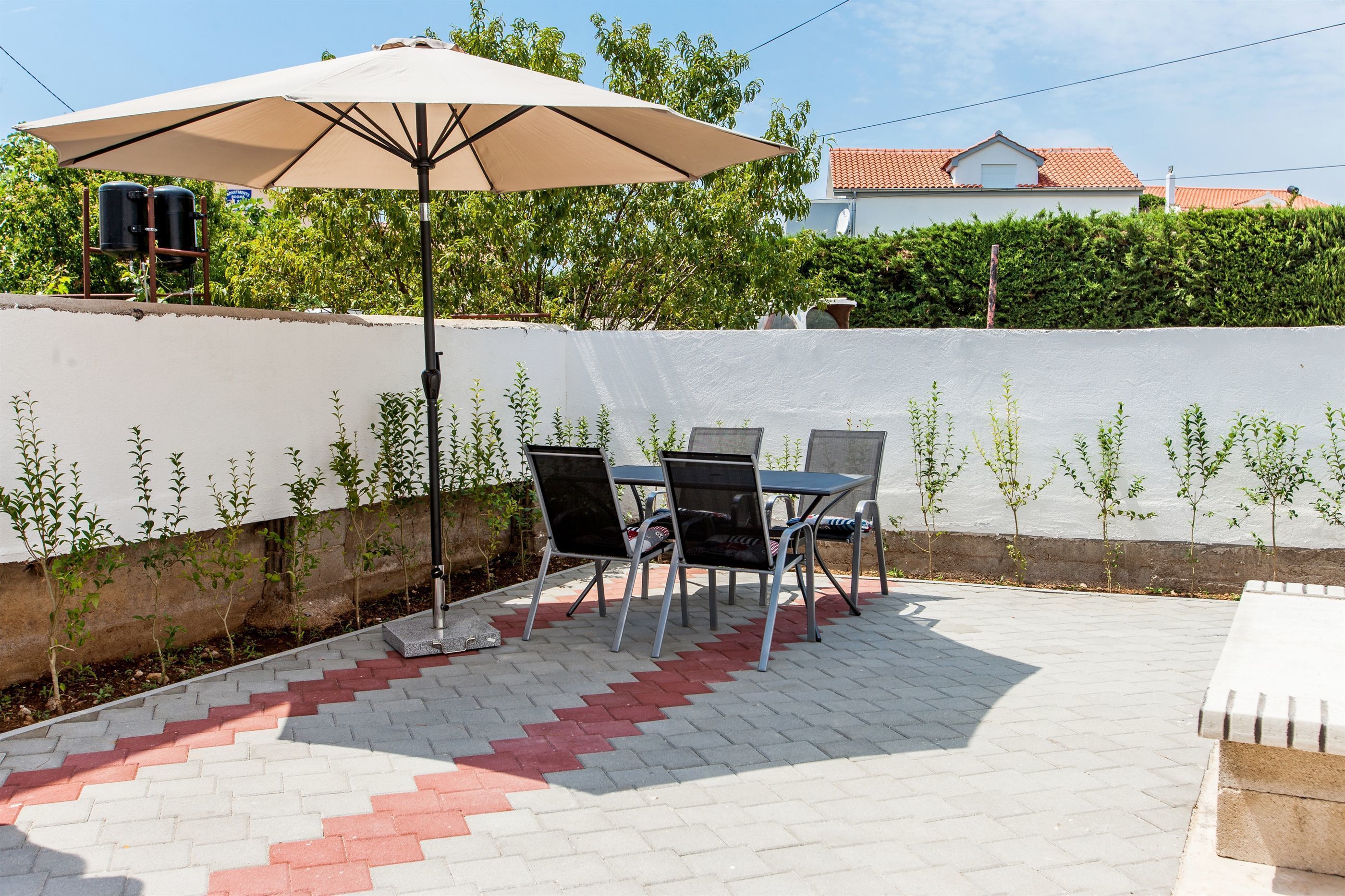 An outdoor patio with a table and chairs under a large umbrella, a white wall with plants, and a building in the background.