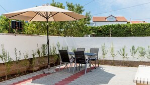 An outdoor patio with a table and chairs under a large umbrella, a white wall with plants, and a building in the background. - House Ivana Tribunj (Tribunj)