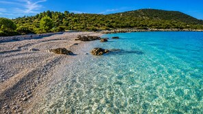 A clear beach with pebbles and rocks, a calm sea, and a green hill in the background. - Apartments Barica (Primosten)