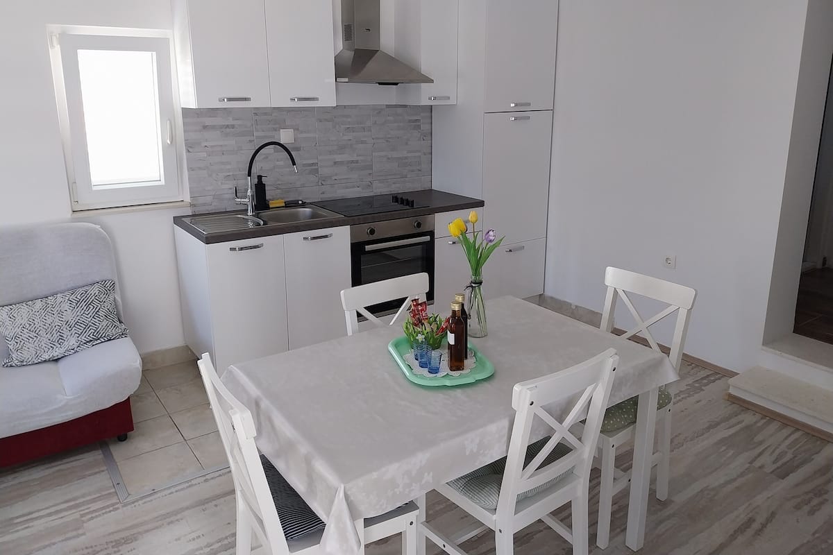A modern kitchen with white cabinets, a black countertop, and a built-in oven. There is a dining table with white chairs and a vase of flowers.