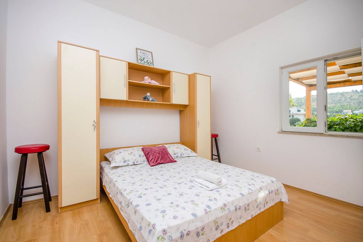A bedroom with a bed, a red stool, a wooden wardrobe, and a window with a view of greenery.