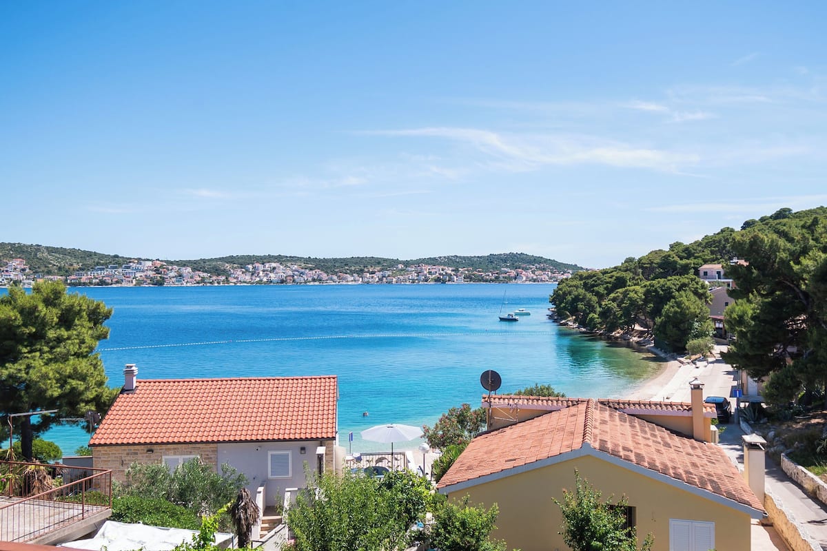 A coastal town with a clear blue sea, houses with red-tiled roofs, and a view of the bay.