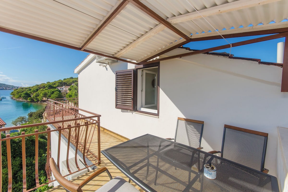 A balcony with a table and chairs, overlooking a coastal area with trees and a body of water.