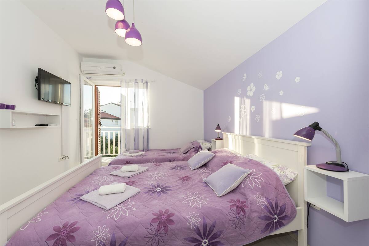 A bedroom with two beds, purple floral bedding, a TV, a bedside table with a lamp, and a window with curtains.