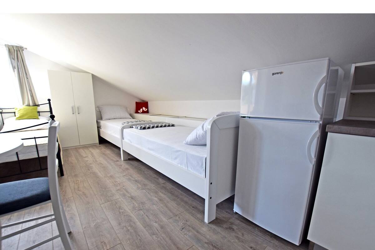Une petite pièce propre, avec un lit simple, une armoire blanche, un bureau avec une chaise et un réfrigérateur blanc.