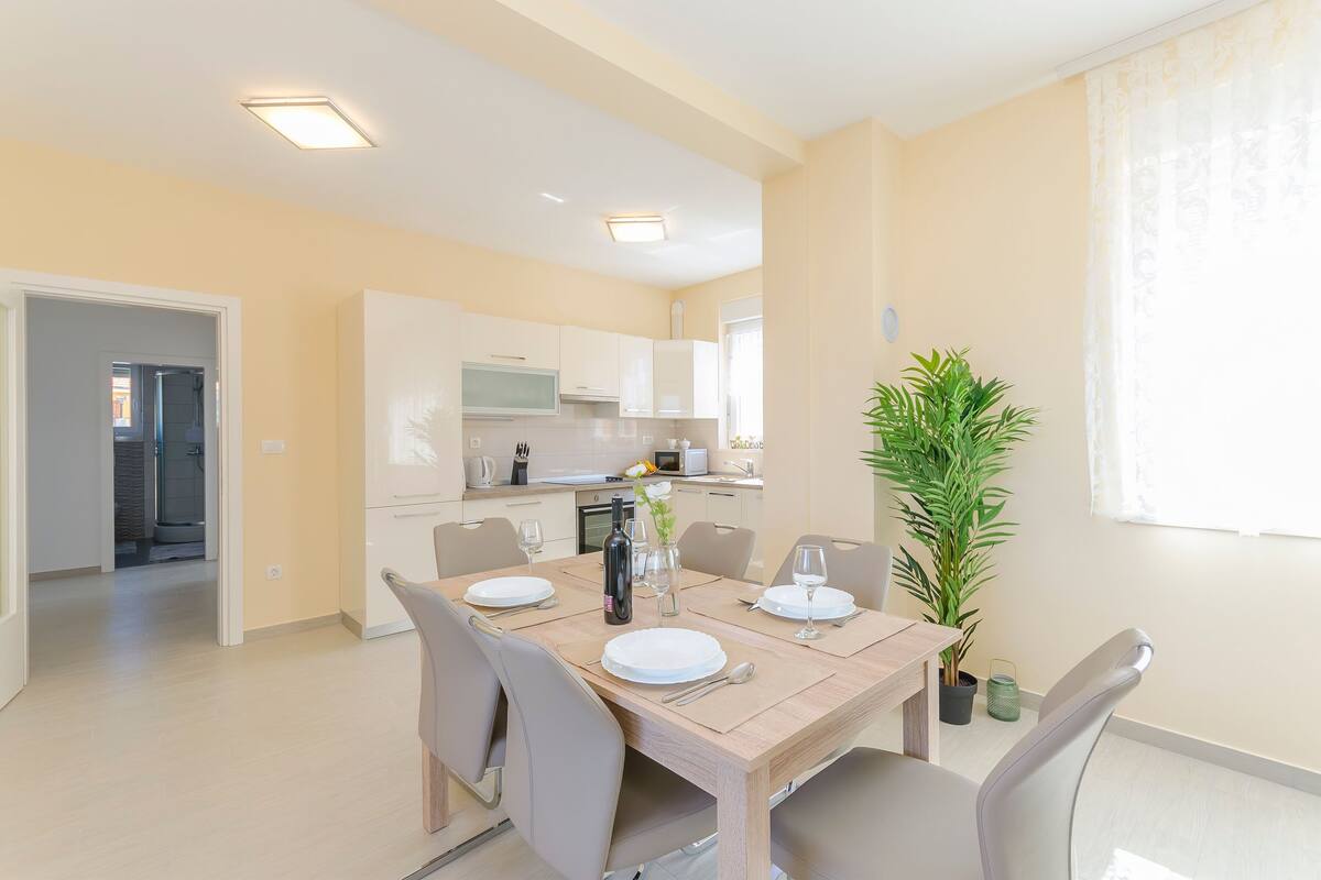 A modern dining area with a wooden table set for four, surrounded by chairs. A potted plant is on the table, and the room has a neutral color scheme with light-colored walls and ceiling.