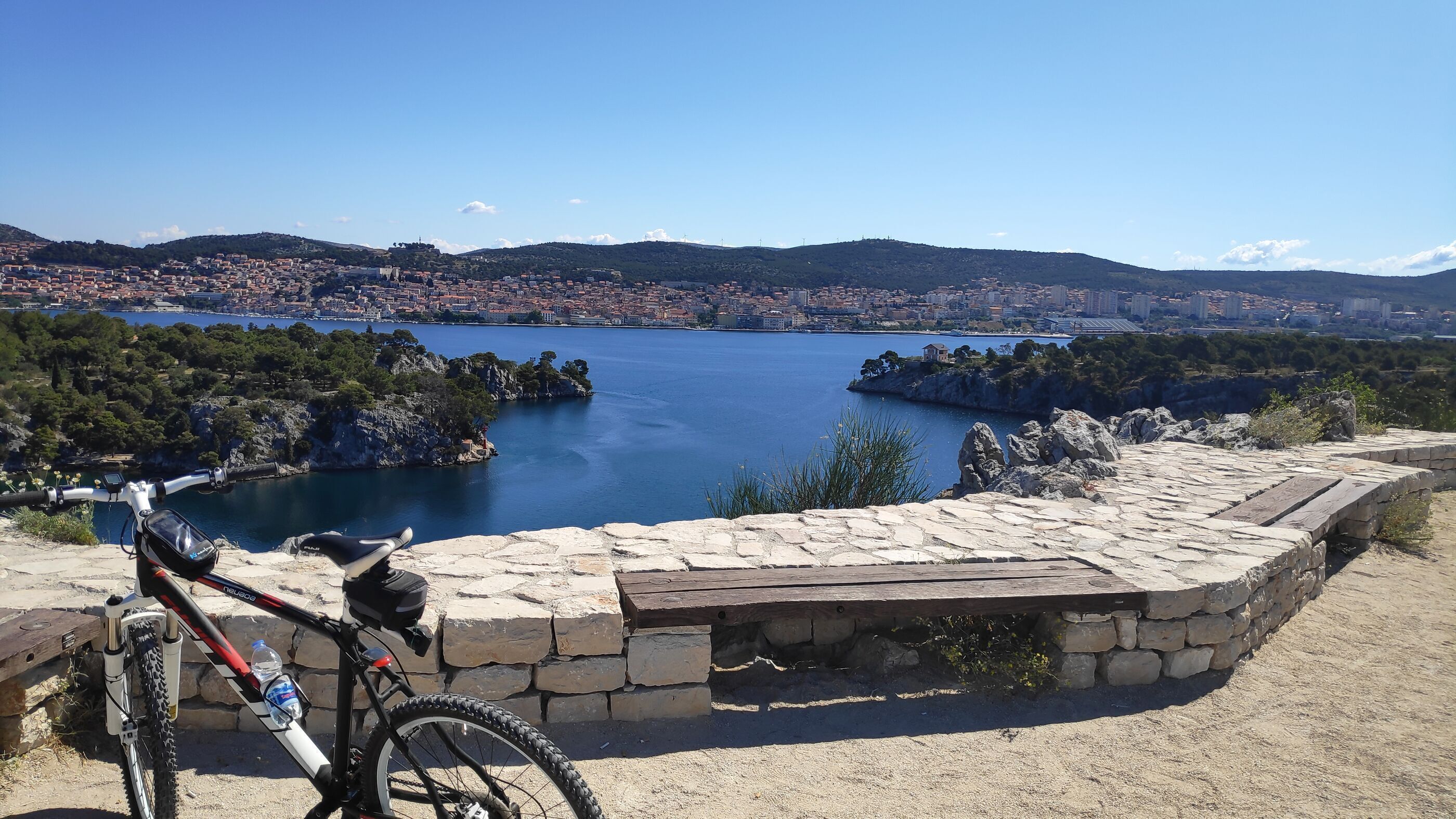 Una bicicleta de montaña estacionada en un saliente de piedra con vistas a un lago y una ciudad al fondo.