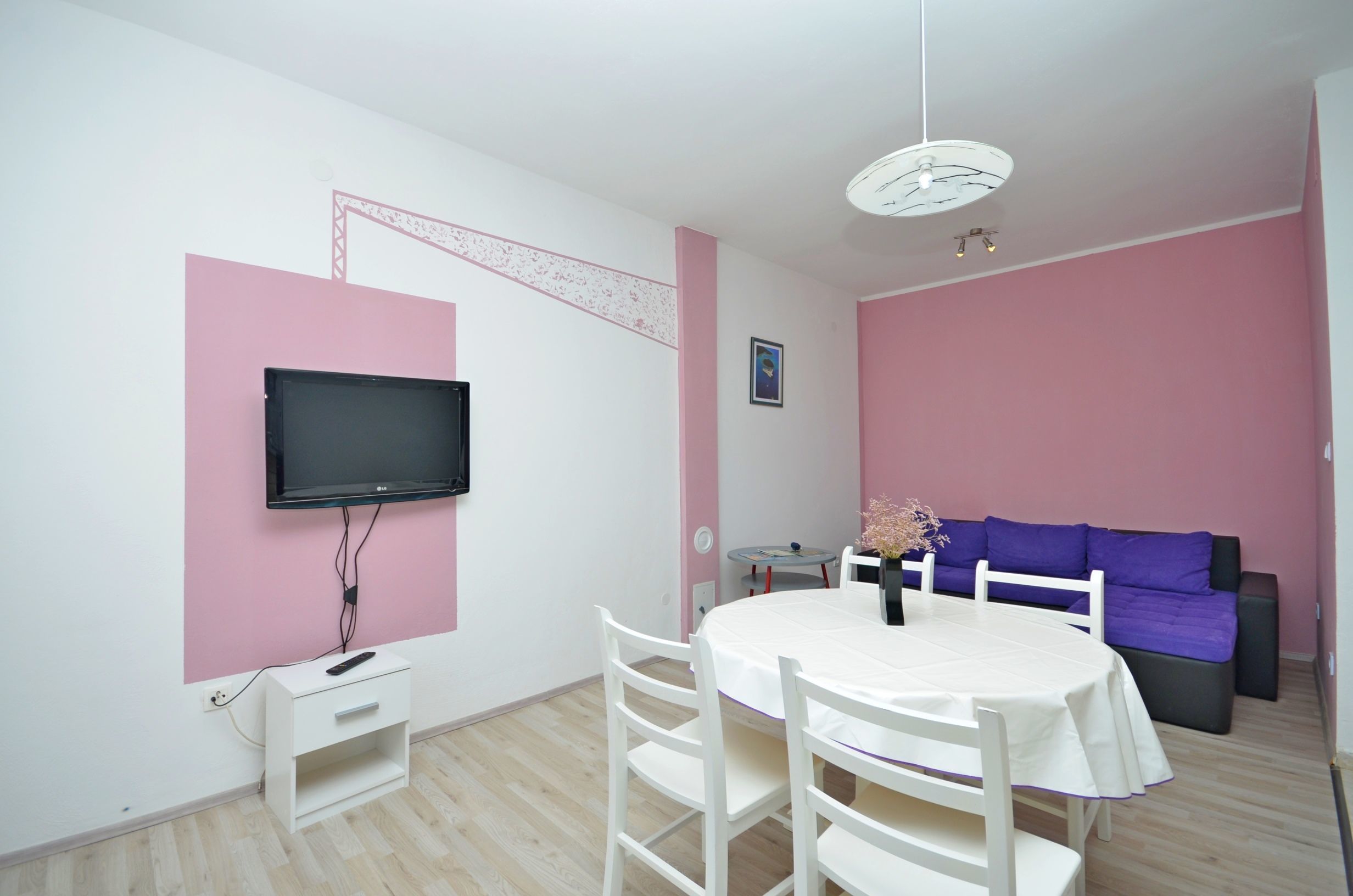 A room with a TV mounted on the wall, a round table with chairs, a purple sofa, and a white cabinet.