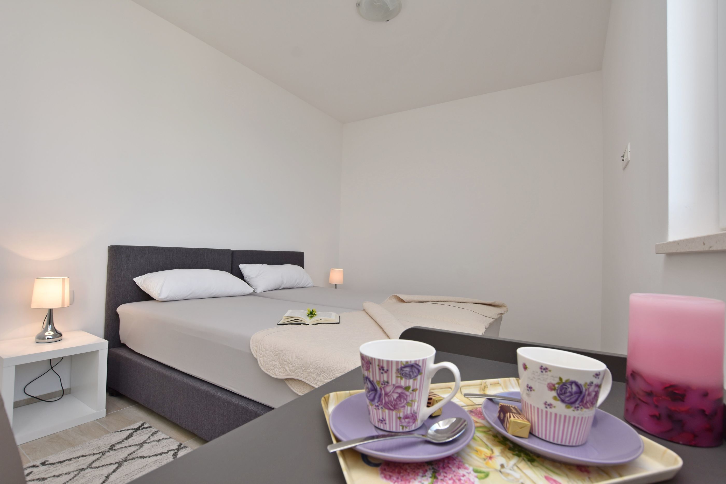 A modern hotel room with a bed, bedside table, lamp, and a breakfast tray with a mug and a teapot.