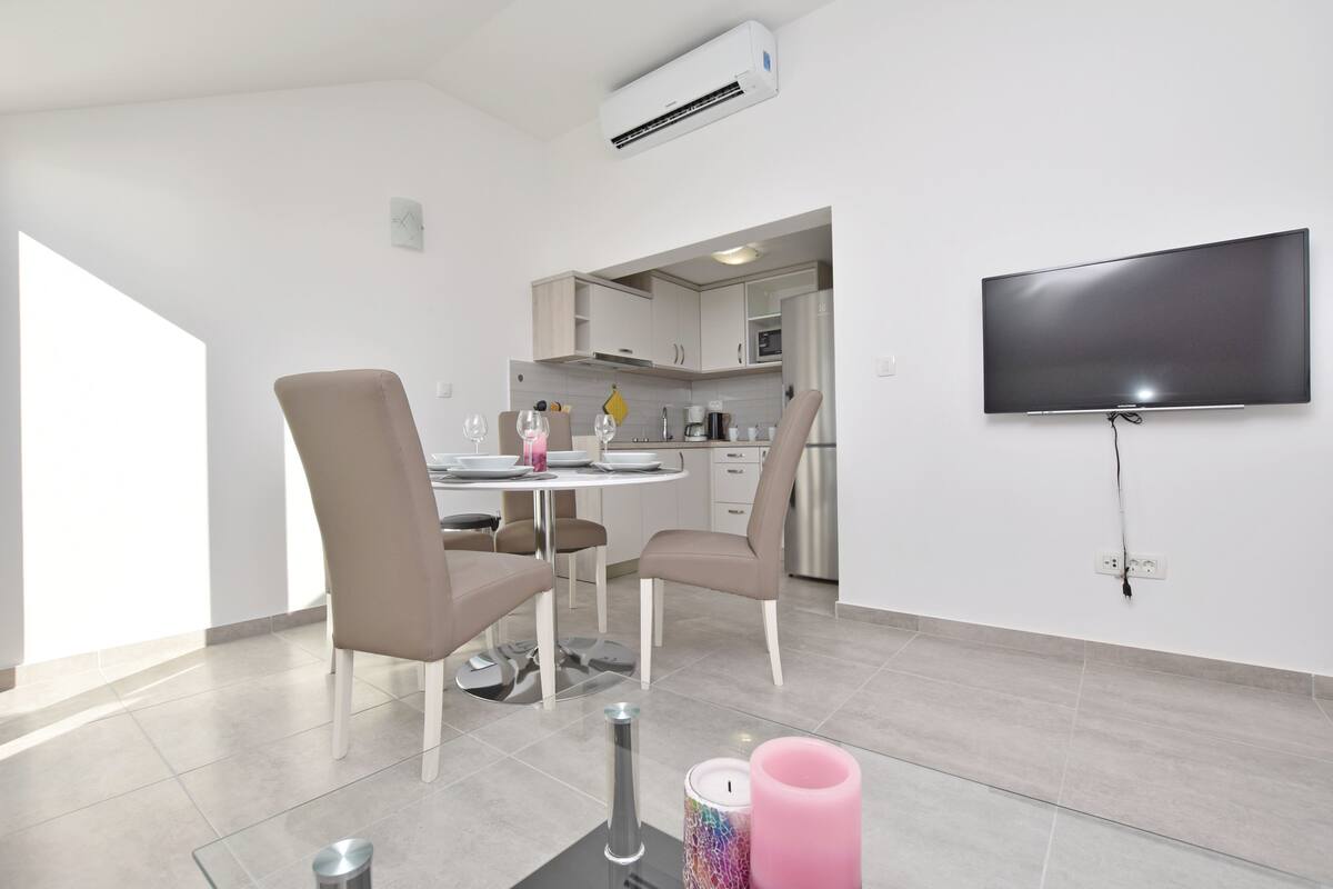 A modern kitchen with a dining area, a flat-screen TV mounted on the wall, and a small trash bin on the floor.