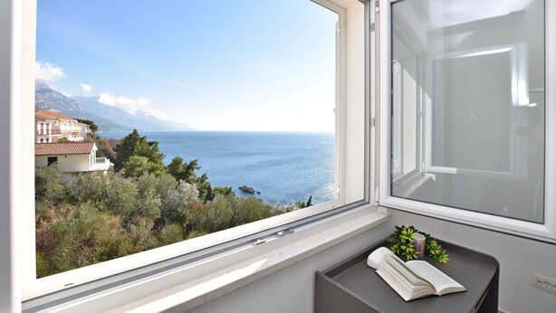 A room with a large window overlooking a coastal view, a small table with a book, and a potted plant. - Apartments Danolic (Pisak)