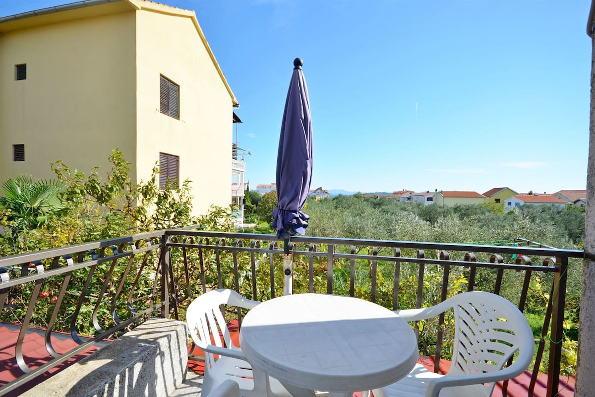 A balcony with a table and chairs, a statue, and a view of a yellow building and greenery.