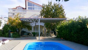 Una piscina con zona coperta, circondata da un giardino e una casa con balcone.