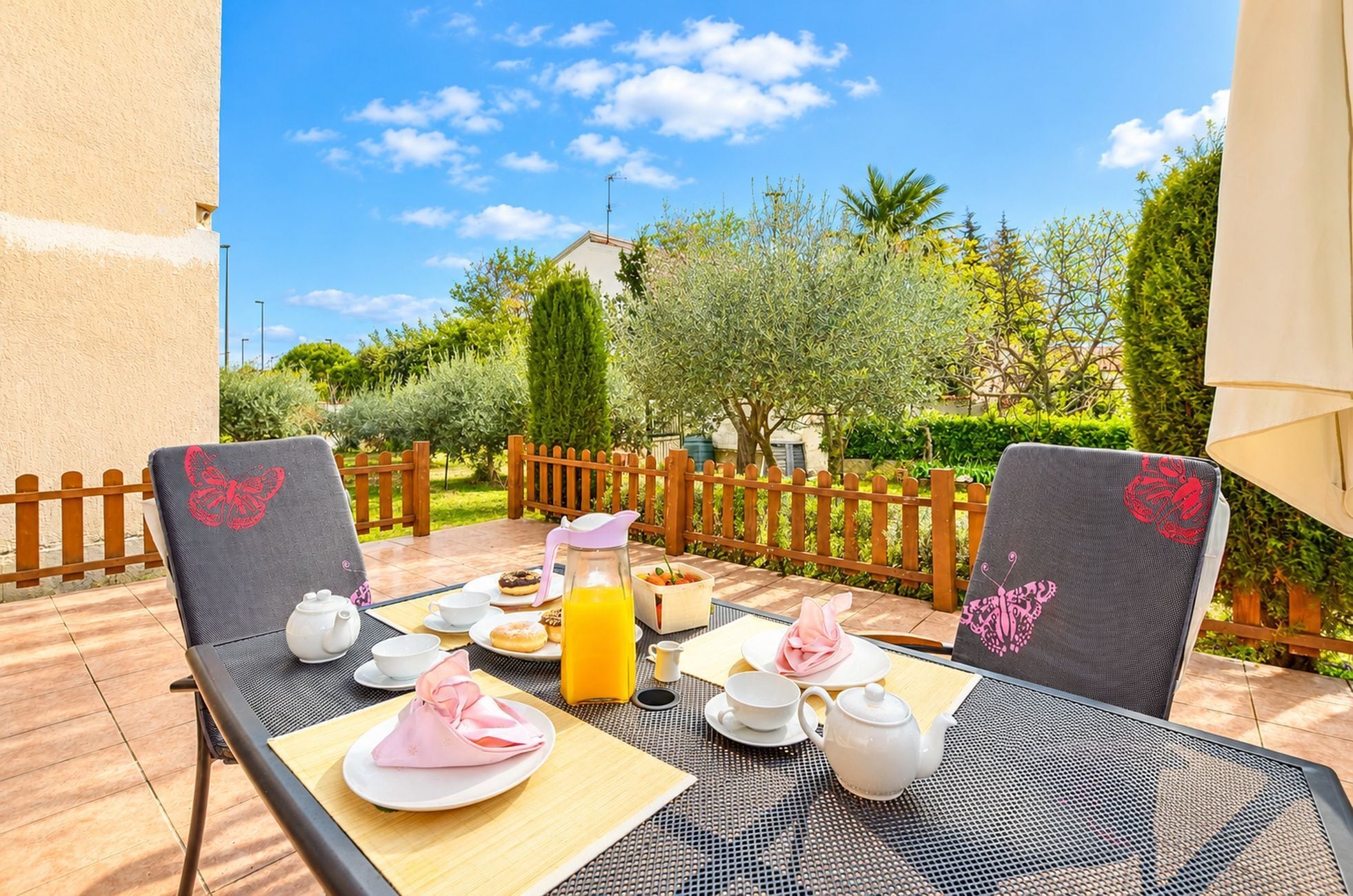 An outdoor dining area with a table set for breakfast, including a teapot, cups, and a fruit bowl.