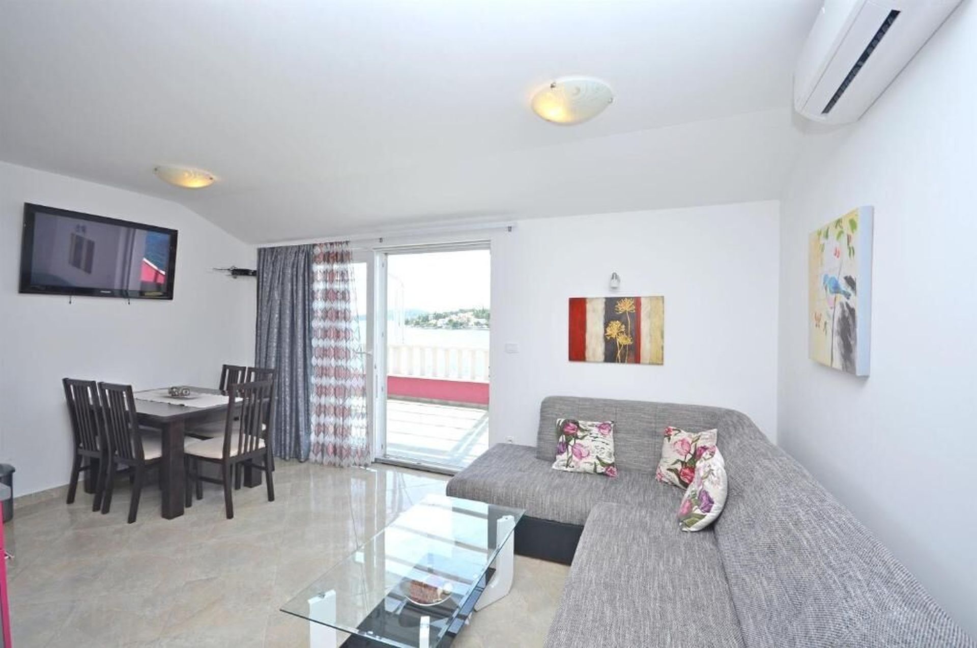 A modern living room with a sofa, a glass coffee table, a dining area with chairs, and a TV mounted on the wall.