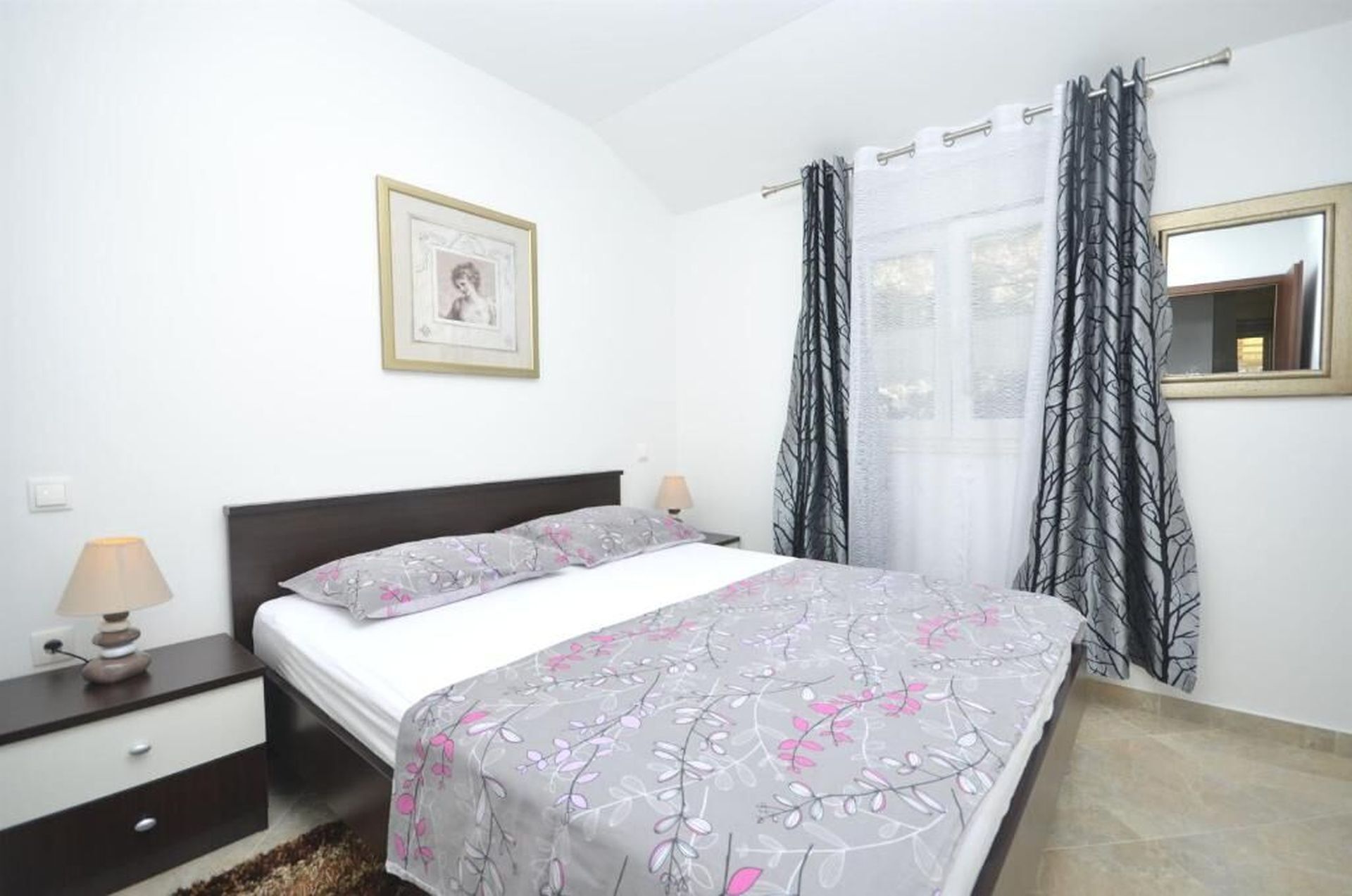 Un dormitorio con cama, mesitas de noche, lámpara, un cuadro enmarcado y una ventana con cortinas.