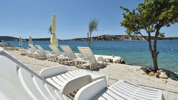 Un área de descanso junto a la playa con sillones blancos y sombrillas, una playa de guijarros y vistas al mar y colinas lejanas.