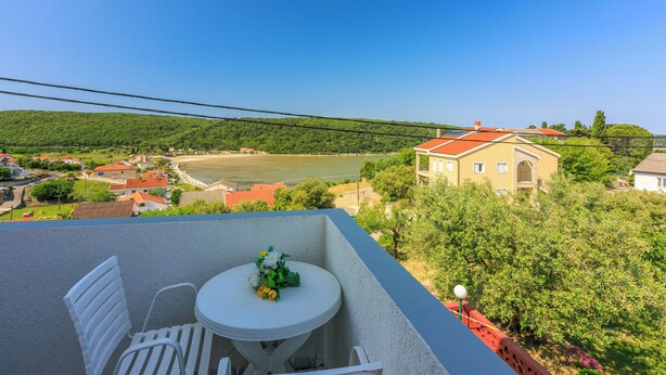 A balcony with a table and chairs, overlooking a river and a hillside with buildings. - Apartments Carmen (Kampor)