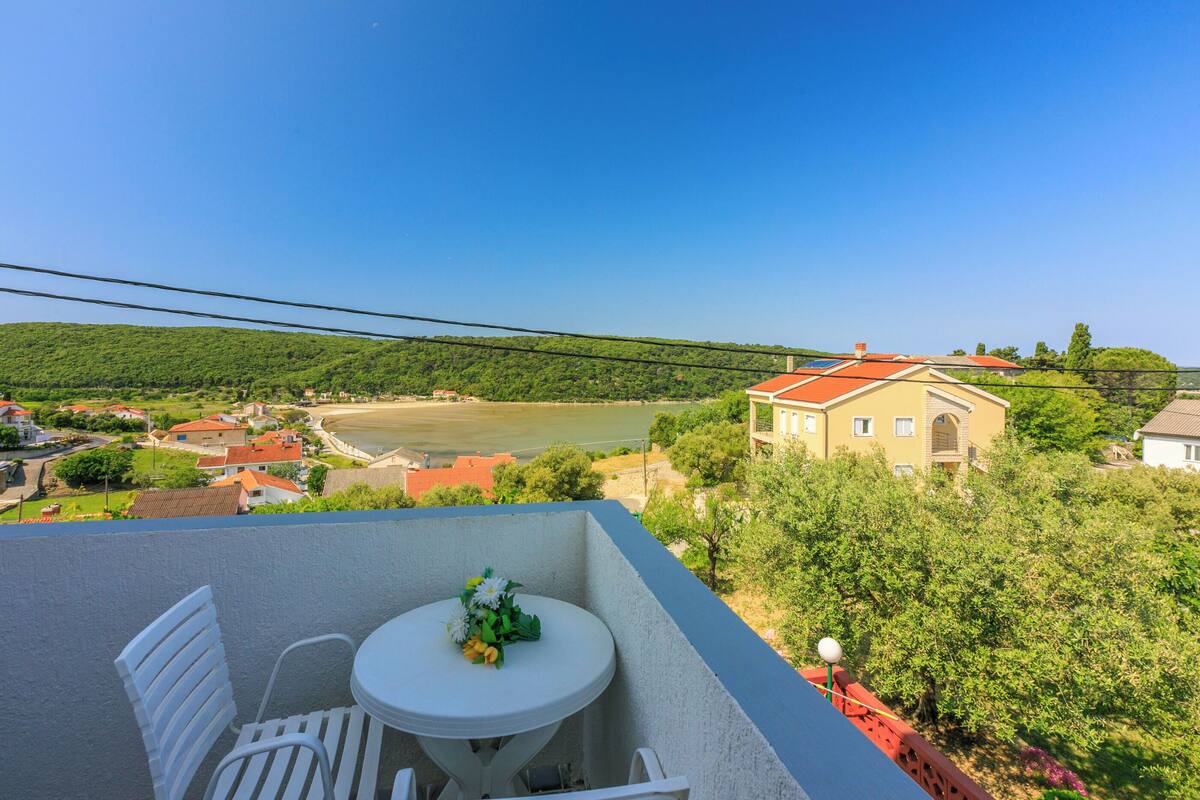 A balcony with a table and chairs, overlooking a river and a hillside with buildings.