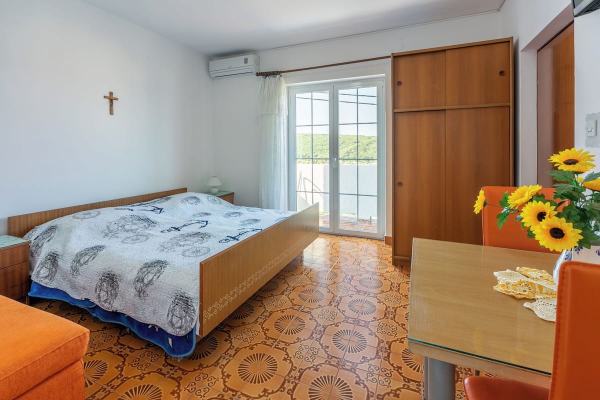 A bedroom with a bed, a table with sunflowers, a cross on the wall, and a sliding door to the balcony.