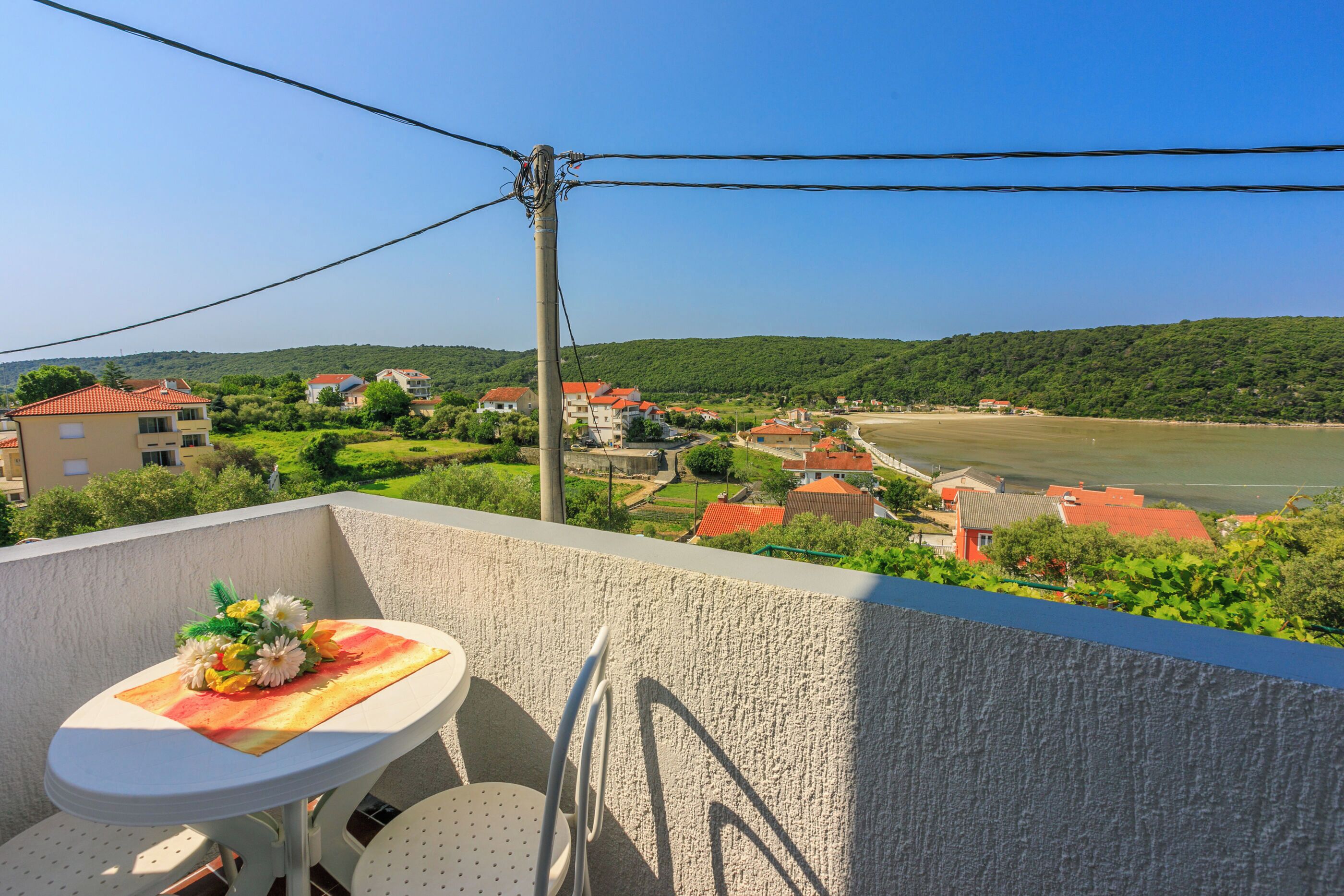 Ein Balkon mit Tisch und Stühlen, mit Blick auf einen Fluss und ein Dorf mit roten Dächern.