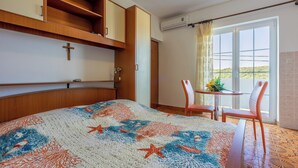 A hotel room with a bed, a small table with chairs, a cross on the wall, and a window with a view. - Apartments Carmen (Kampor)