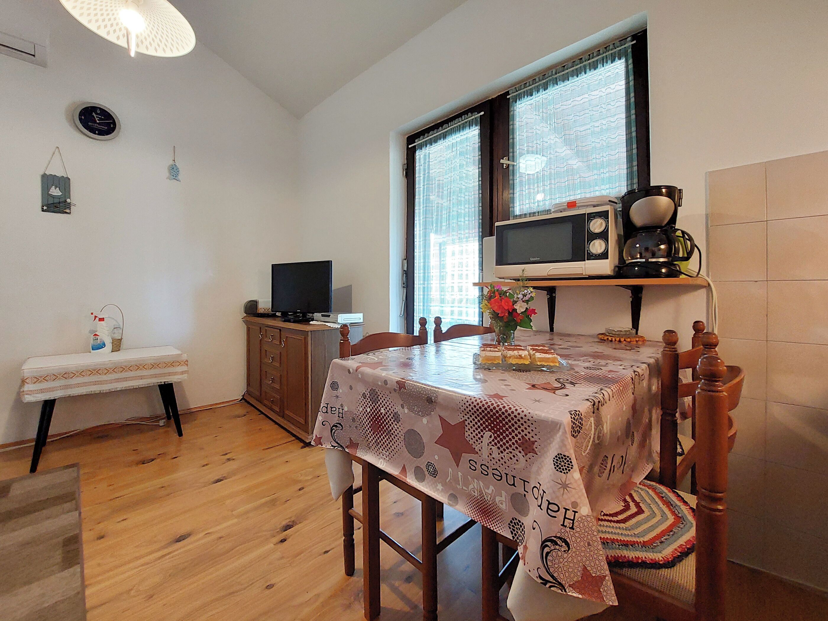 A dining area with a table set for two, a television on a cabinet, and a window with curtains.