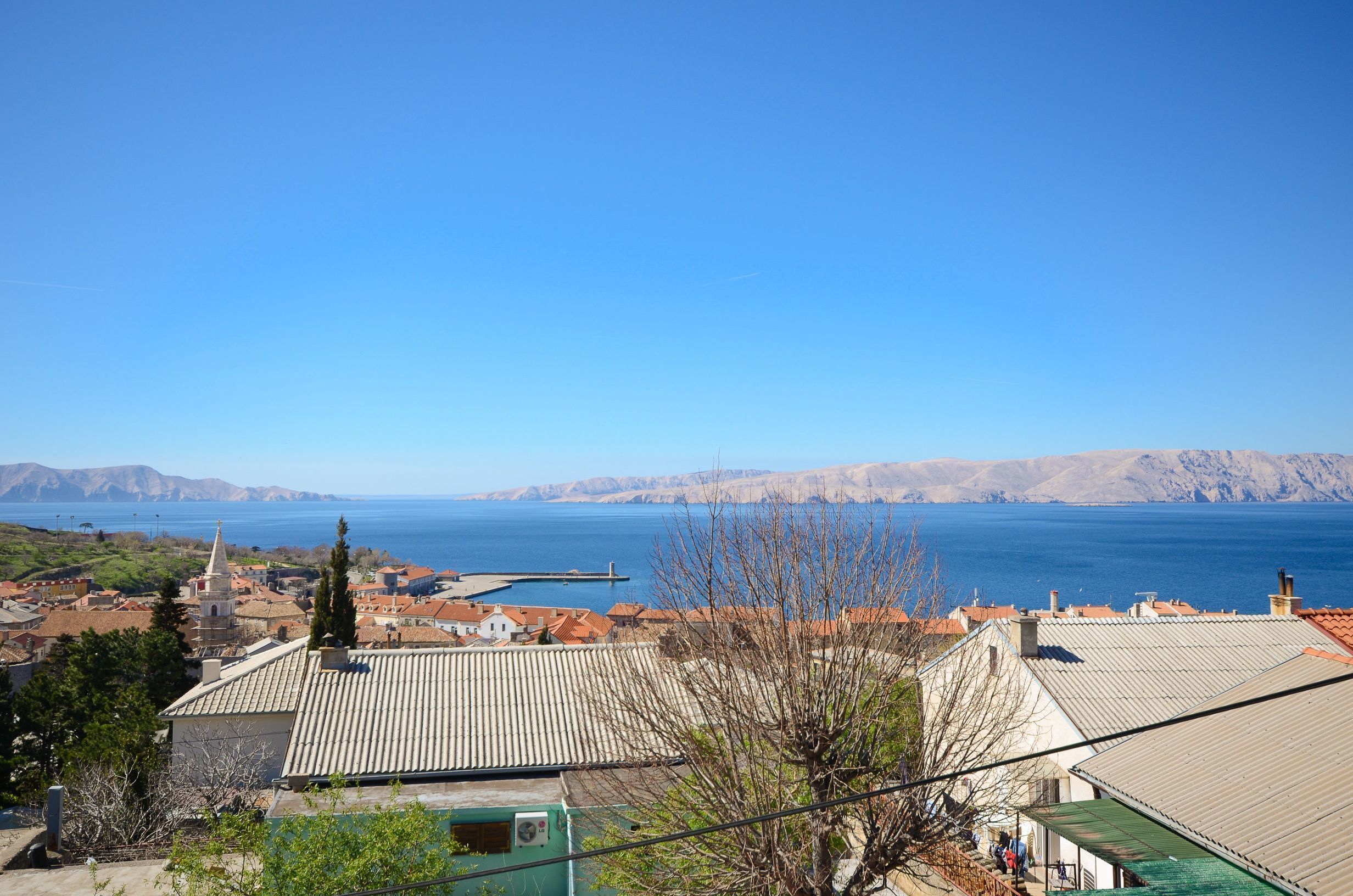 Eine Küstenstadt mit terrakottafarbenen Dächern, blauem Himmel und Blick auf das Meer und ferne Berge.