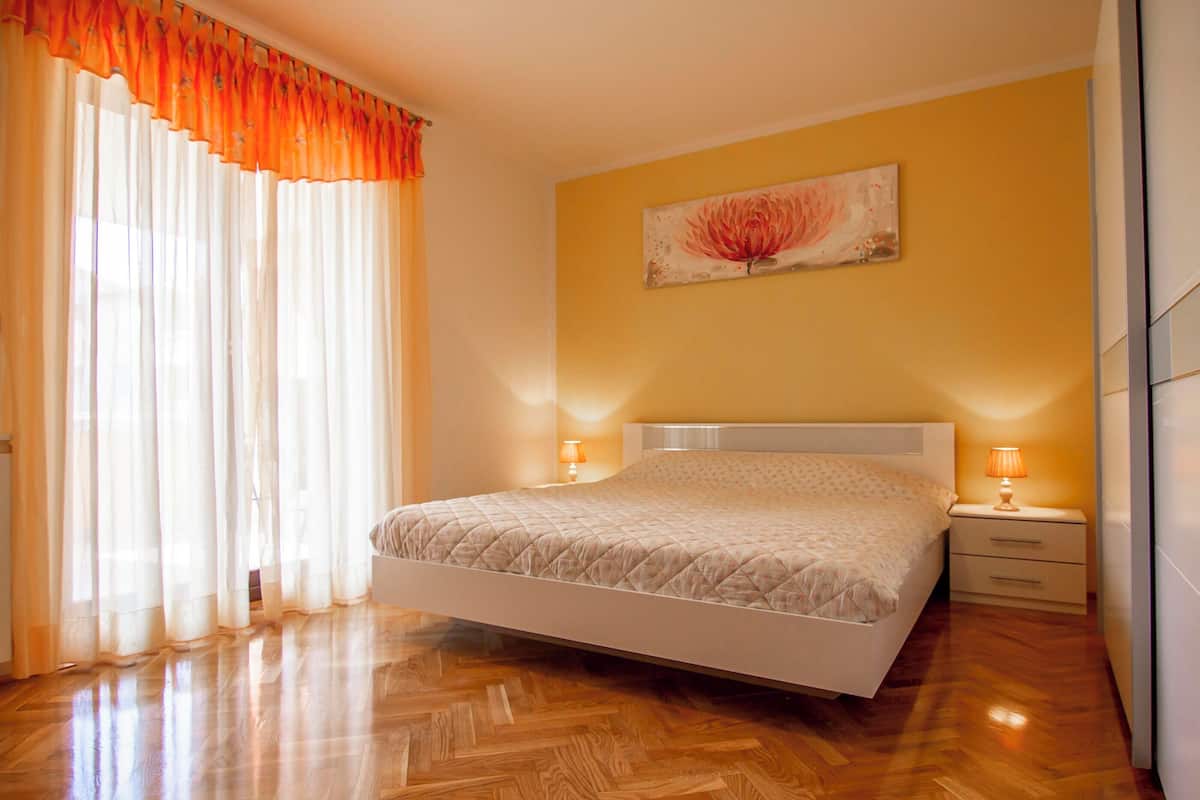A bedroom with a bed, wooden flooring, a nightstand, a lamp, and a wall-mounted painting.