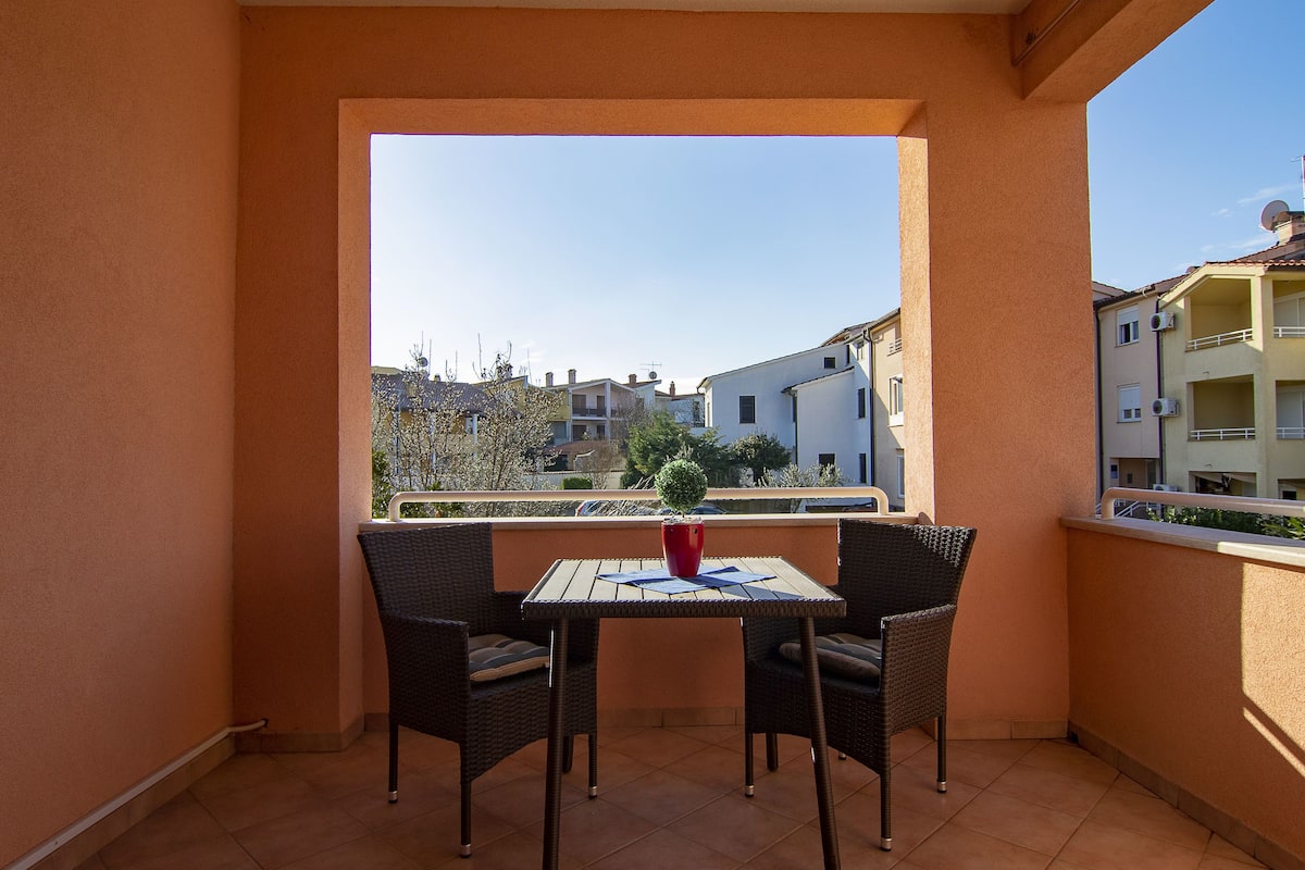 A balcony with a table and two chairs, a potted plant, and a view of neighboring buildings.